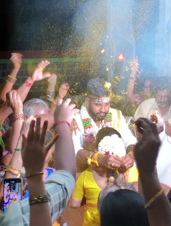 Rajavel and Nivethidha at their wedding, gold confetti falling as family raise their hands around them